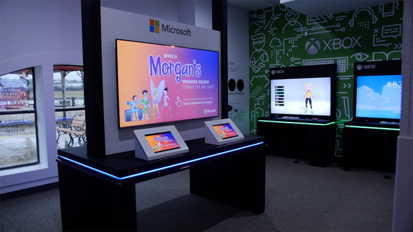An interior space with a promotional setup for Microsoft Xbox. There are two large screens, one mounted on the wall and another on a stand, both displaying colorful graphics related to Xbox. The screen on the wall has text that reads “Which Morgan’s world squad character are you?” with cartoon characters displayed below the text. The screen on the stand shows a single character from the game. Below this screen, there is a platform with blue LED lighting and two interactive tablets placed in front of it for users to engage with the content being displayed. The background wall is decorated with green graphics featuring gaming-related icons and symbols, creating an immersive environment that highlights Microsoft’s gaming brand.
